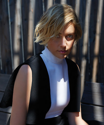 Portrait of a blonde woman with short hair wearing a white top and black dress, posed in an outdoor setting with a wood background.