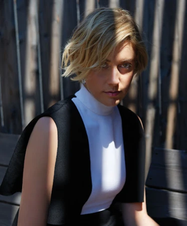 Portrait of a blonde woman with short hair wearing a white top and black dress, posed in an outdoor setting with a wood background.