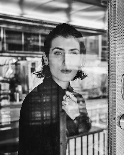 Black-and-white portrait of a woman in front of a window with reflections, wearing hoop earrings and rings, and looking directly at the camera.
