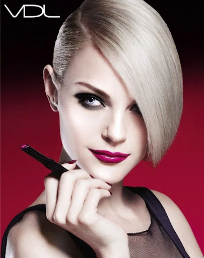 Close-up of a blonde woman with vibrant fuchsia lipstick holding a black cosmetic product, set against a dynamic red gradient background.