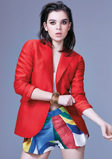 A young woman in a vibrant red blazer and colorful patterned shorts poses against a grey backdrop in a studio fashion shot.