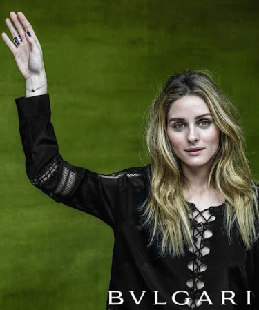 Portrait of Olivia Palermo with long blonde hair wearing a black lace-up top and raising her right arm against a green background