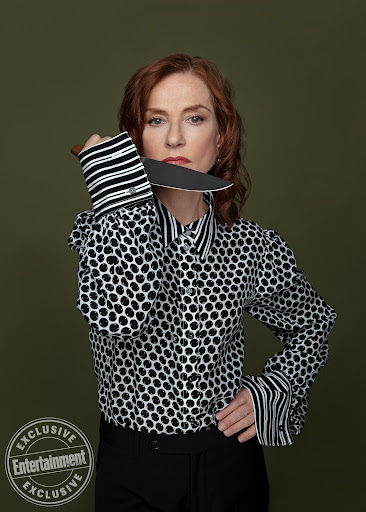 Portrait of a Caucasian woman holding a knife to her neck, wearing a black and white polka dot blouse, against an olive background.
