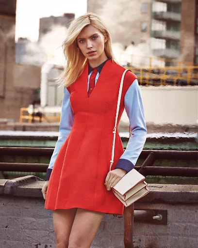 Blonde woman in a red dress and blue long-sleeved shirt stands on a rooftop with buildings in the background, holding a cream-colored purse.