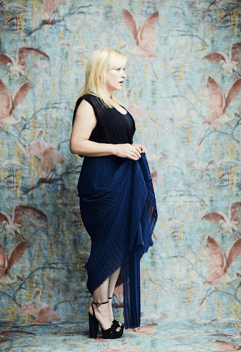 Woman posing in black top and pleated blue dress with black platform heels in front of a floral and bird patterned wallpaper.