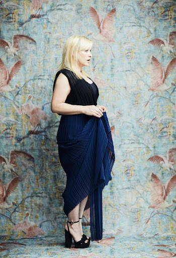 Woman posing in black top and pleated blue dress with black platform heels in front of a floral and bird patterned wallpaper.