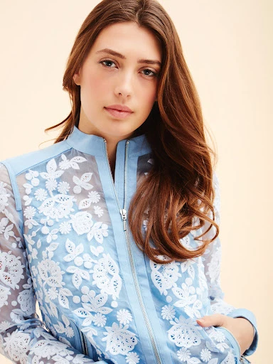 Close-up studio shot of a brunette adult woman posing in a baby-blue floral lace jacket with a zipper, in a neutral environment.