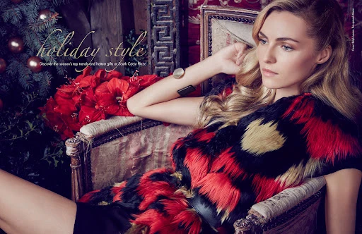 A fashion editorial shot of a blonde woman wearing a red, black, and gold fur coat, posing in an ornate chair near holiday decor.