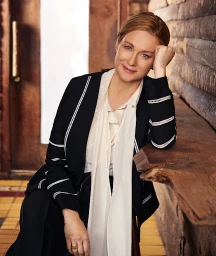 Portrait of a woman with red hair wearing a black jacket with white trim and a white silk blouse, resting her head on her hand.