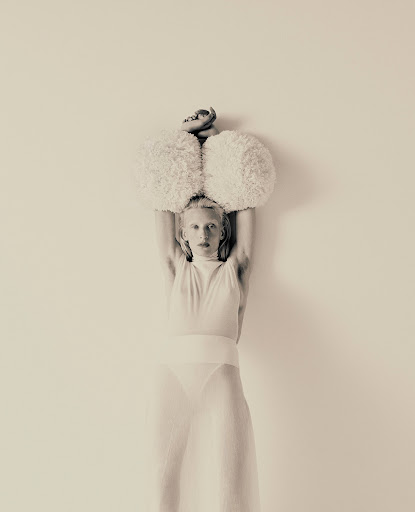 Artistic studio shot of an adult woman in a sheer white outfit, holding fuzzy white balls above her head against a solid backdrop.