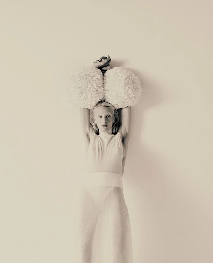Artistic studio shot of an adult woman in a sheer white outfit, holding fuzzy white balls above her head against a solid backdrop.