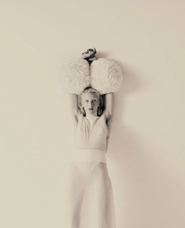 Artistic studio shot of an adult woman in a sheer white outfit, holding fuzzy white balls above her head against a solid backdrop.