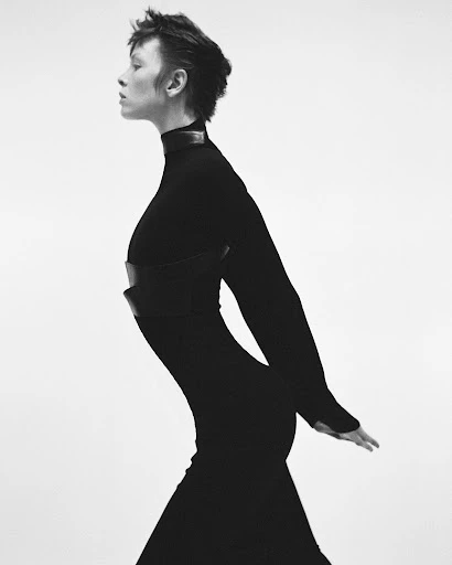 Elegant studio shot of a slender woman in a long black dress with a leather corset, set against a clean white backdrop, profile pose.