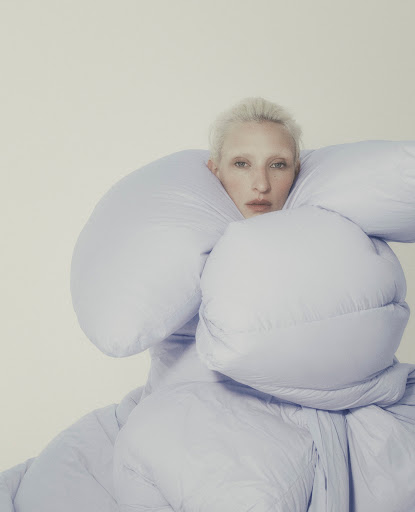Fashion portrait of a woman with platinum blonde hair and pale skin wearing a light purple, puffy down jacket against a white background.