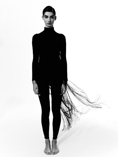 Monochrome studio portrait of a slender adult female in a black turtleneck and leggings, standing against a stark white backdrop.