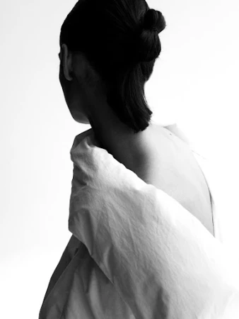 Close-up of a model in profile with dark hair tied in a bun, wearing a white garment draped over bare shoulders against a white background.