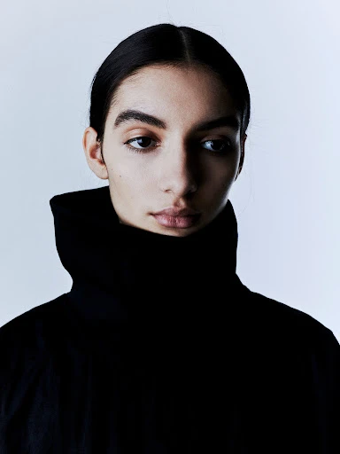 Close-up portrait of a young adult woman with dark hair wearing a high-collared black top against a bright, neutral backdrop in a studio setting.