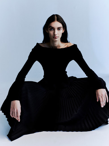 Elegant woman in a black, long-sleeved dress poses in a minimal studio setting with a smooth, light blue-grey background.