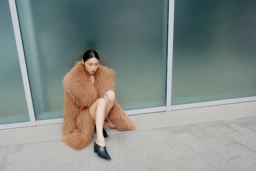 Asian female model with neutral expression seated in oversized fur coat and high-heeled mules, posing in front of frosted glass panels.