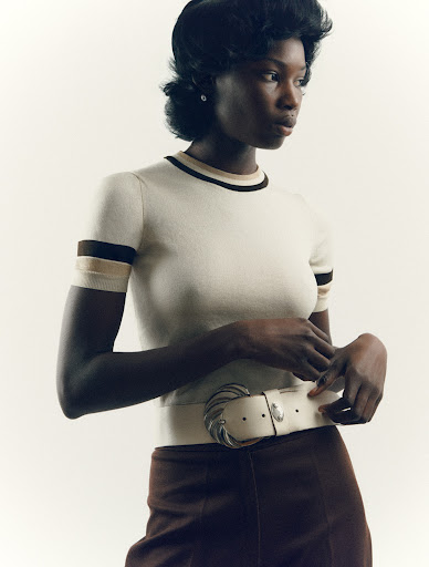 Elegant studio portrait of a Black woman in a cream top with brown trim and dark trousers, posing against a neutral backdrop.
