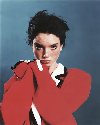 Fashion portrait of a woman with dark hair in a red jacket against a blue background, arms crossed, looking directly at the viewer.