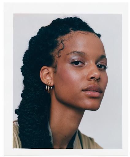 Close-up portrait of a young Black woman with intricate braids, gold hoop earrings, and a nose ring against a neutral background.
