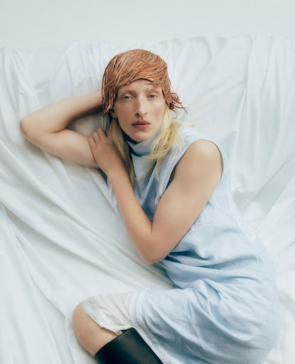 Woman lying on white fabric wearing a blue sleeveless dress and black boots, with wet blonde and brown hair in a studio setting.