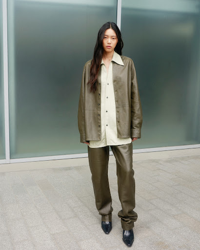 A woman with long dark hair poses in an olive leather jacket and trousers in an outdoor fashion shot against a frosted glass wall.