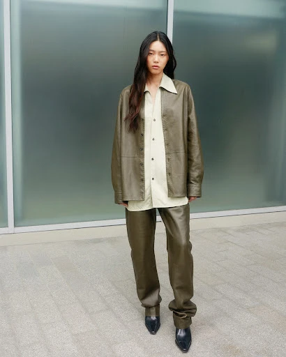 A woman with long dark hair poses in an olive leather jacket and trousers in an outdoor fashion shot against a frosted glass wall.