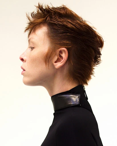 Profile shot of an adult female with short auburn hair wearing a black turtleneck and a wide black collar against a white background.