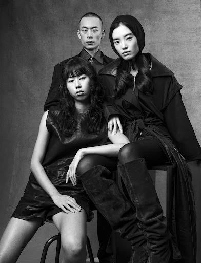 Black and white studio portrait of three Asian adults in modern dark clothing. One man and two women, one seated on a stool. Fashion photography.