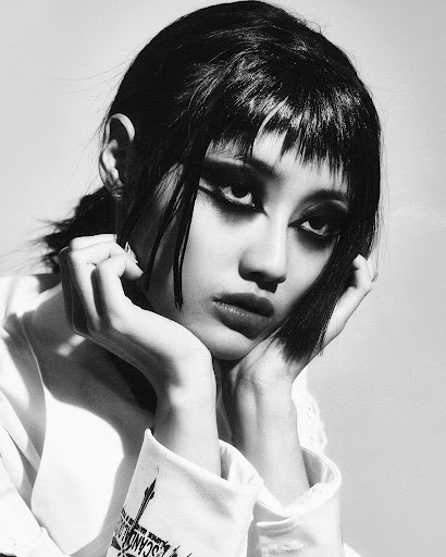 Monochrome portrait of an adult woman with blunt bangs and dramatic makeup, holding her hands under her chin in a close-up.