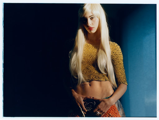 Portrait of a young woman with fair skin and blonde hair wearing a gold crop top, ornate belt, and a neutral expression.