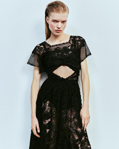 Model with slicked-back hair wearing a black lace cut-out dress against a pale blue seamless backdrop.