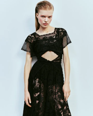 Model with slicked-back hair wearing a black lace cut-out dress against a pale blue seamless backdrop.