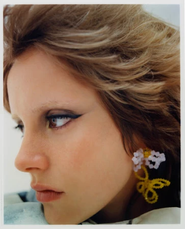 Close-up portrait of a woman with dramatic winged eyeliner and beaded flower earrings against a plain background