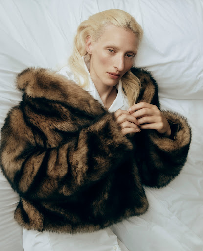 Fashion shot of a fair-skinned adult female with blond hair, wearing a fur coat, lying on a white bed sheet in a studio setting.