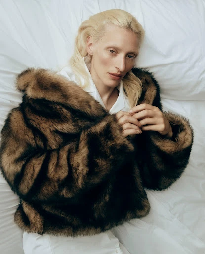 Fashion shot of a fair-skinned adult female with blond hair, wearing a fur coat, lying on a white bed sheet in a studio setting.