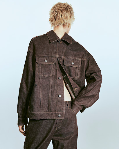 Person in a dark brown denim jacket and pants, shot from the back, posing against a light blue gradient background in a studio.
