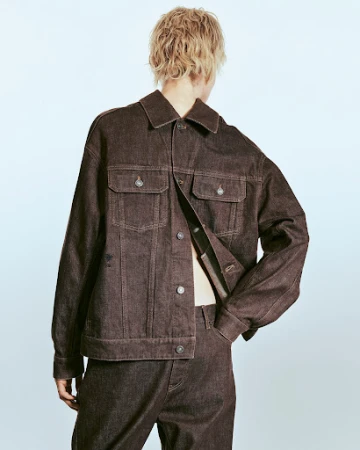 Person in a dark brown denim jacket and pants, shot from the back, posing against a light blue gradient background in a studio.