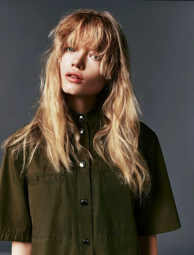 A fair-skinned young woman with blonde wavy hair and bangs poses in a studio wearing an olive green button-down shirt with short sleeves.