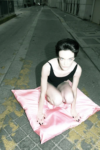 A young woman in a black bodysuit kneels on a pink pillow in an urban alleyway, looking directly at the camera in an unusual pose.