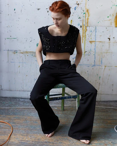 Redhead woman in a black crop top and black wide-leg pants sits on a blue stool in a photo studio with an artfully painted backdrop.