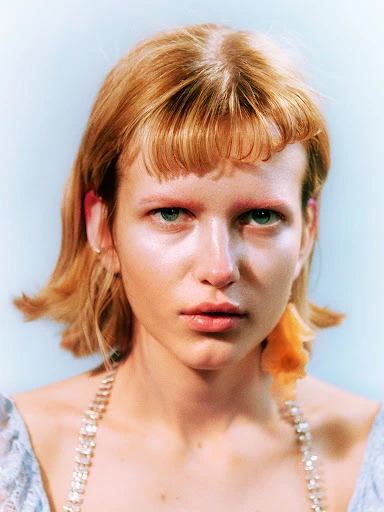Portrait of a blonde woman in a blue floral top and rhinestone necklace with a light blue background, captured in a studio setting.