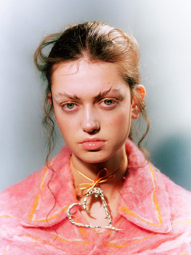 Close-up portrait of a young woman with exaggerated eyebrows wearing a pink jacket and a heart-shaped necklace on a pale background.