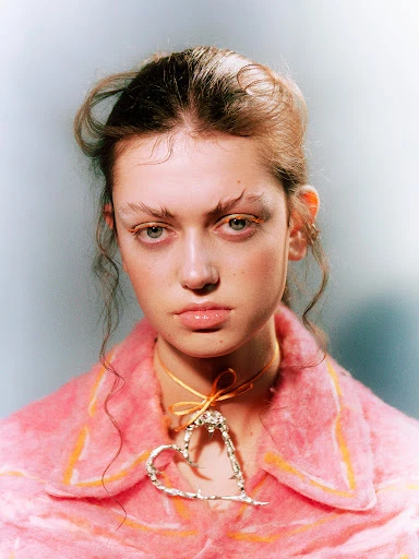 Close-up portrait of a young woman with exaggerated eyebrows wearing a pink jacket and a heart-shaped necklace on a pale background.