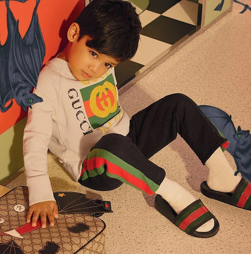 Young child poses indoors wearing Gucci sweatshirt and slides, with patterned wall and floor, and bat and umbrella artwork.