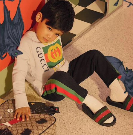 Young child poses indoors wearing Gucci sweatshirt and slides, with patterned wall and floor, and bat and umbrella artwork.