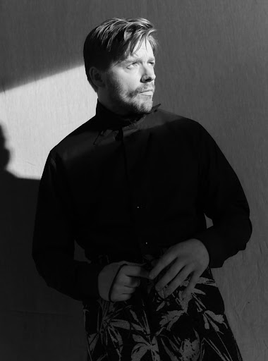 Black and white portrait of a man with fair hair and a beard, wearing a dark shirt and patterned trousers, leaning against a wall.