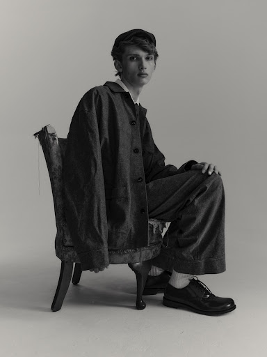 Monochrome portrait of a young adult male model wearing oversized workwear, seated on a distressed antique chair against a neutral backdrop.